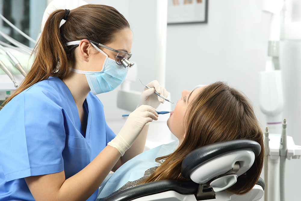 Dentist wearing eyeglasses gloves and mask examining a patient teeth with a dental probe and a mirror in a clinic box with equipment in the background