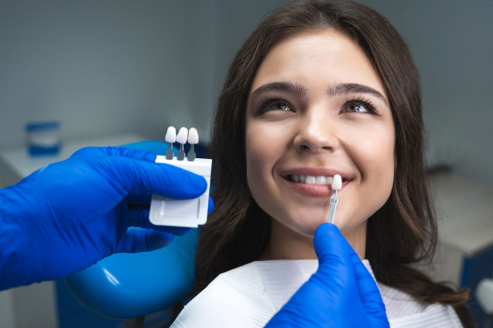 dentist in blue medical gloves applying sample from tooth enamel scale to happy woman patient teeth to pick up right shade for teeth bleaching procedure.
